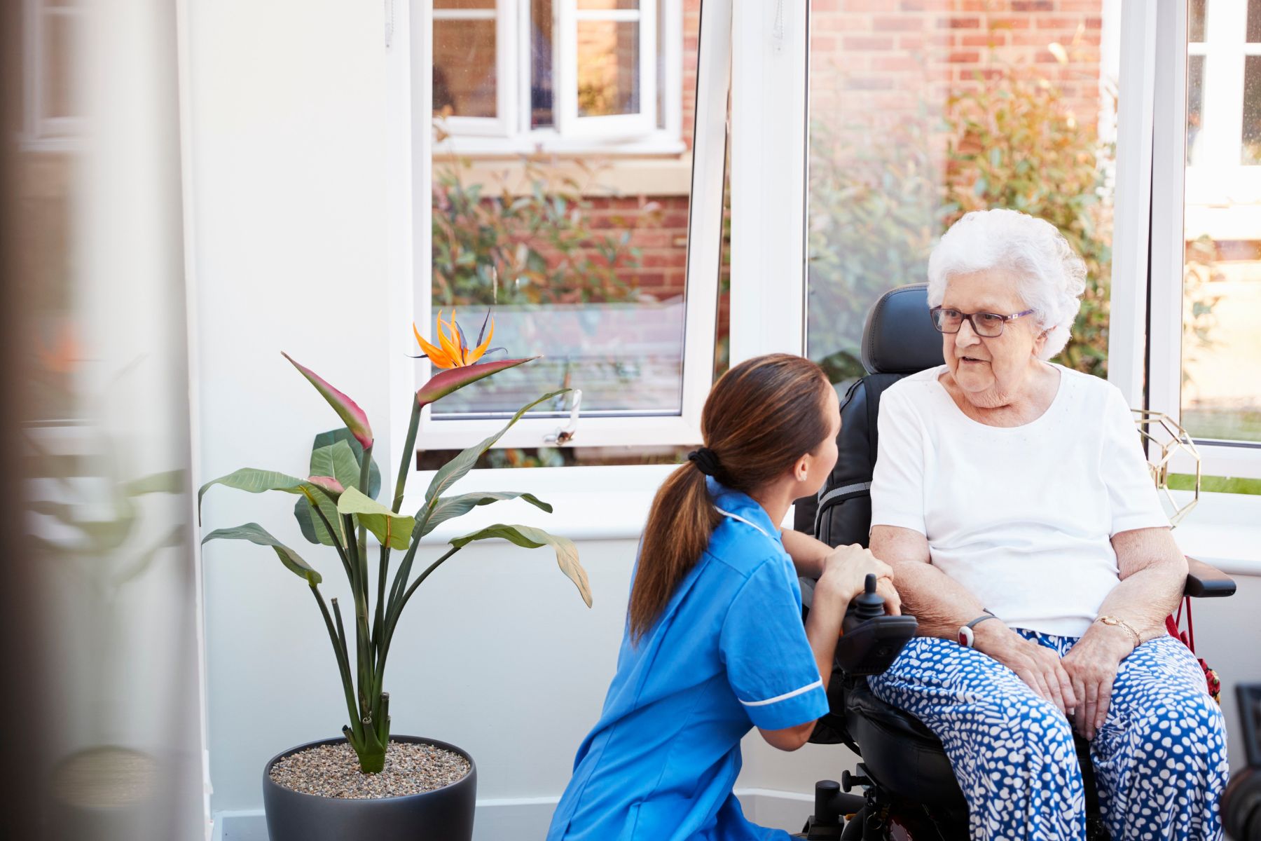 Senior Woman Sitting In Motorized Wheelchair Talking With Nurse In Retirement Home